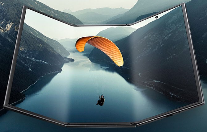 Samsung Galaxy Tri Fold phone shown from the front with the sides partially folded in. On the screen you see a paraglider over a body of water and you see the same images on the phone's screen.