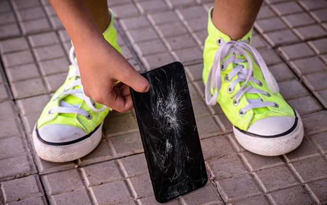 Hand picking up phone with broken display from brick surface with neon green sneakers.